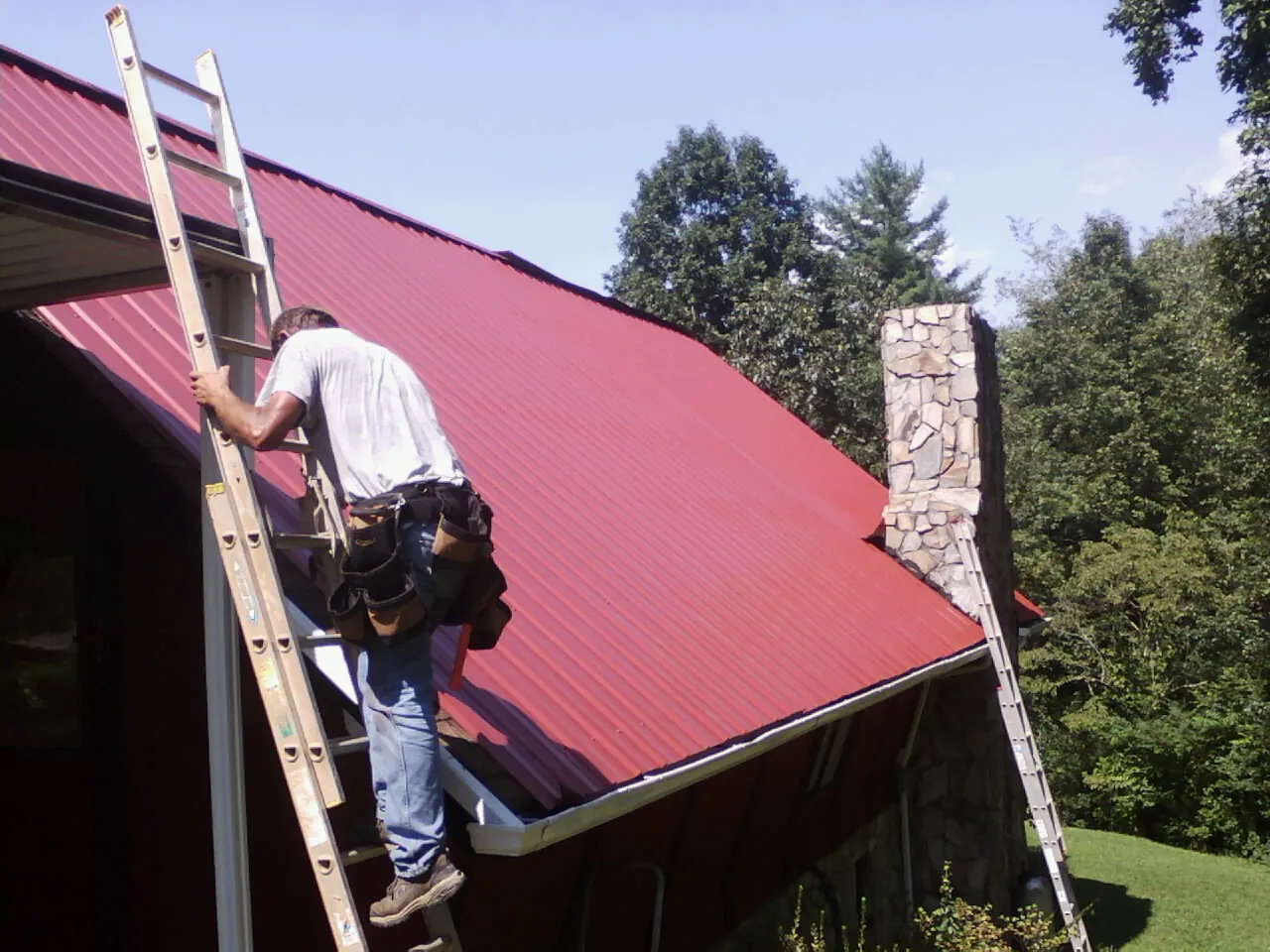 Roofer on a ladder beside a red metal roof and stone chimney — Soffit & Fascia Repair by Roofing Brothers in Dennis Port